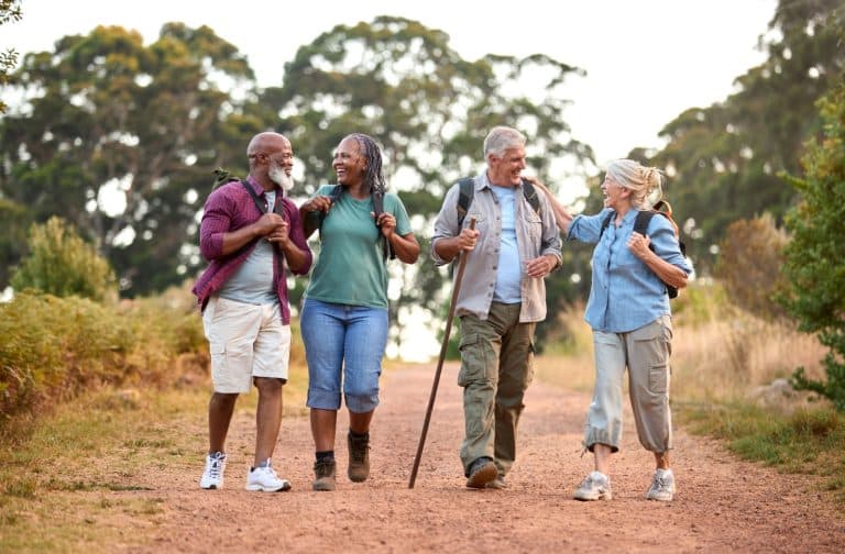 Two older couples laugh together as they walk outside on a dirt trail, the sun shining & big leafy trees surrounding them.