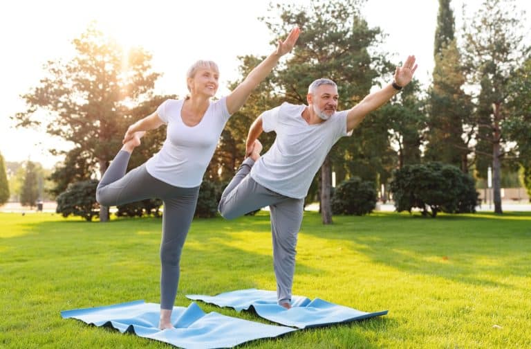 A senior couple performs balance exercises on a sunny, green lawn.