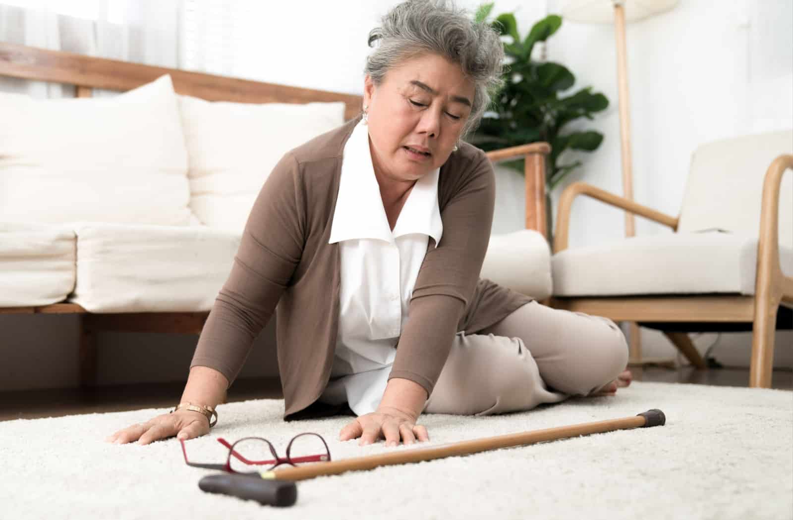 A senior woman fell from the couch. Her mobility aid cane and glasses lay on the carpet in front of her.
