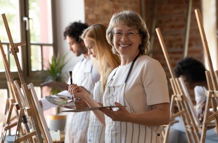 An older adult smiling while holding a paintbrush in front of an easel during a group art activity class.