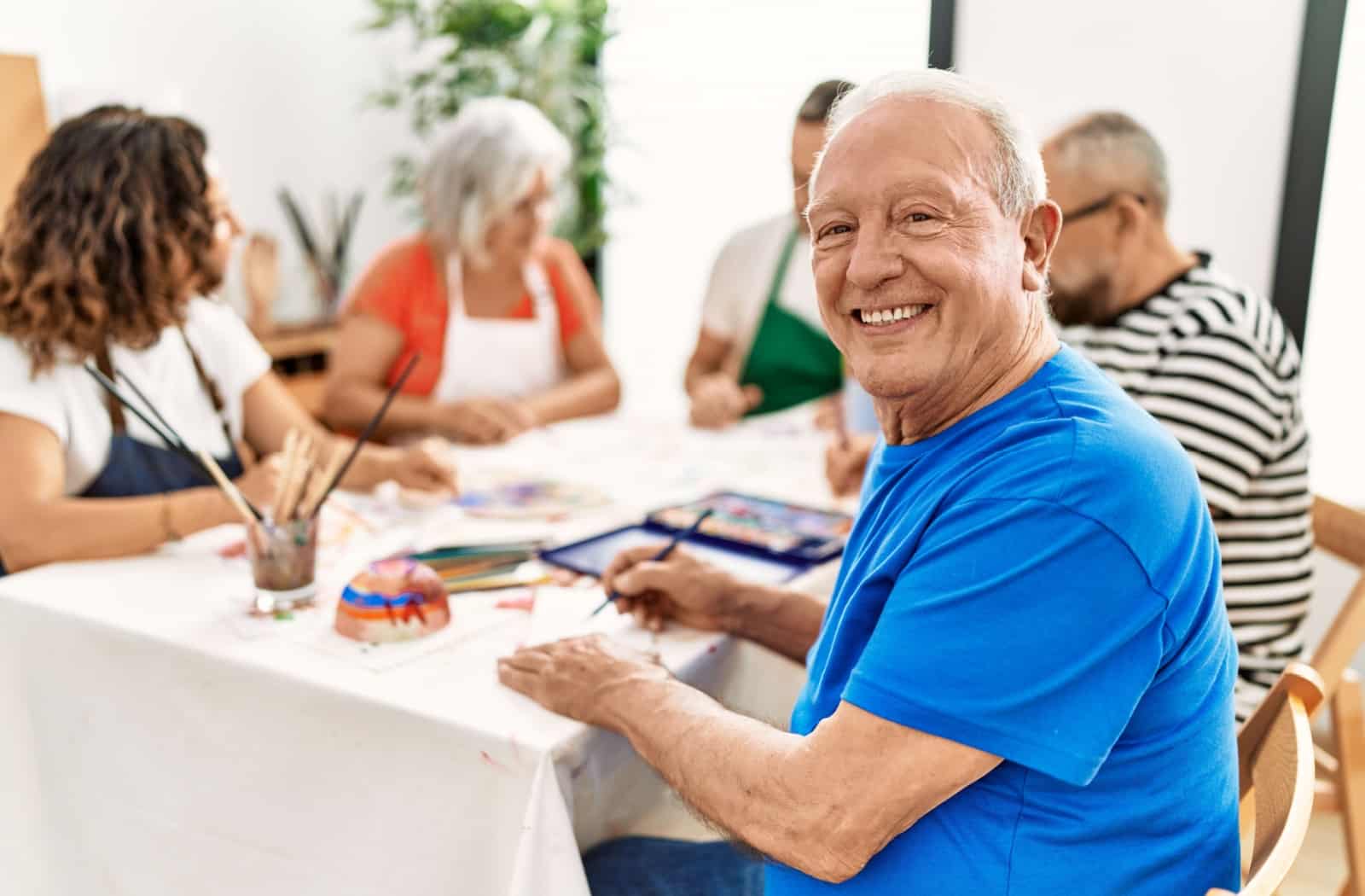 An older adult smiling and looking at the camera while sitting at a table in a group art activity class.
