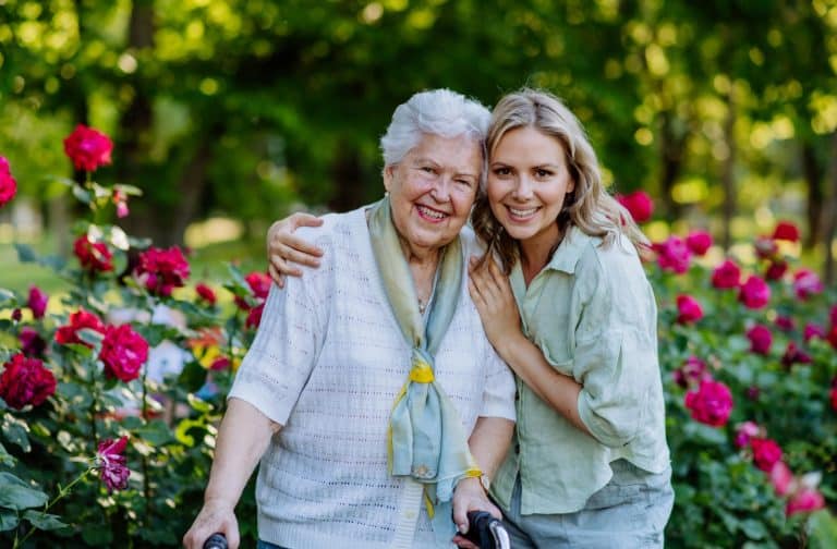 An adult grandchild and grandparent embrace happily in a flower garden.