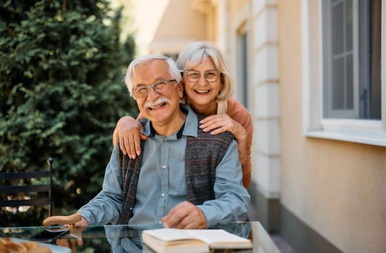 An older person leans on their spouse's shoulder and smiles while outside on a sunny day.