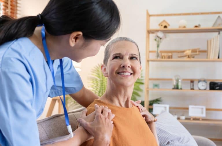 A nurse checks in on a senior resident in their home, holding them by the shoulders as they smile.