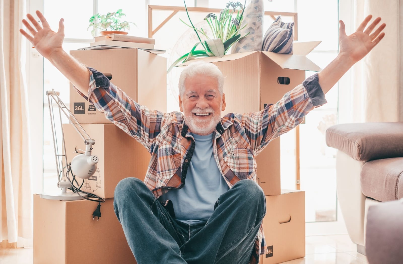 An older individual holds their hands up in celebration as they sit in front of personal items packed in moving boxes.