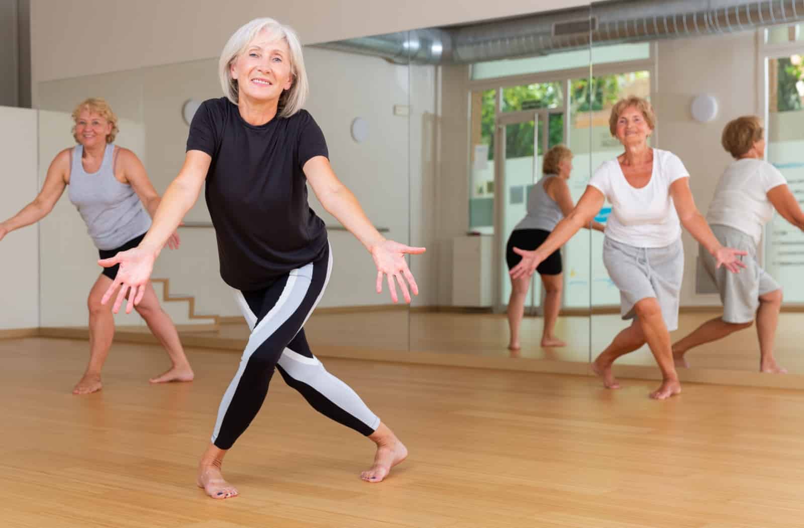 Three smiling seniors are in a dance studio. They lunge and stretch out their hands as part of a dance routine.