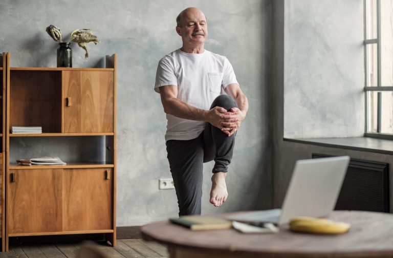 An older adult in their living room in senior living with one knee raised during an exercise to keep their joints healthy.