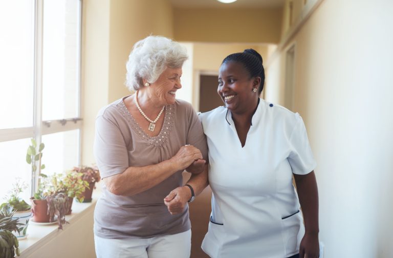 A senior resident and nurse walk arm and arm through the community halls, chatting and smiling.