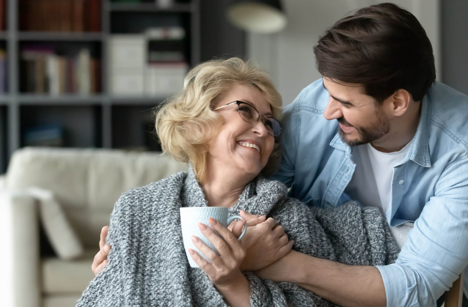 An older adult in senior living sitting on the couch and smiling as their adult child brings them a mug of coffee and hugs them from behind.
