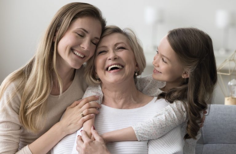 An adult and their young child laughing and hugging, while visiting an older parent in a senior living community.