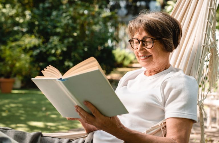 A smiling older adult reads a book outdoors in a senior living community.