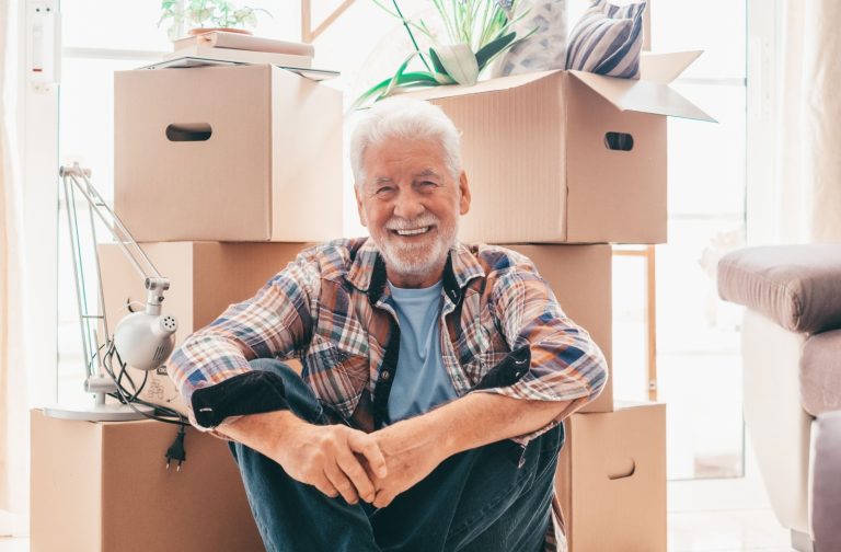 Senior smiling in front of moving boxes, showing a positive and seamless transition during the downsizing process.