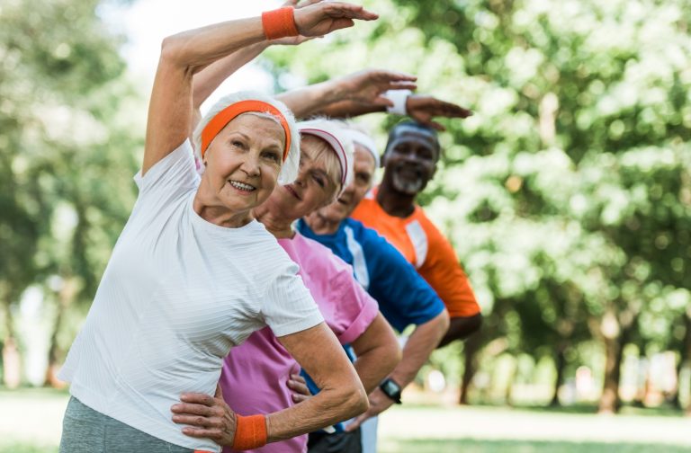 Four seniors, outdoors in athletic wear, stand in a line, each stretching an arm over their head to stretch their hips