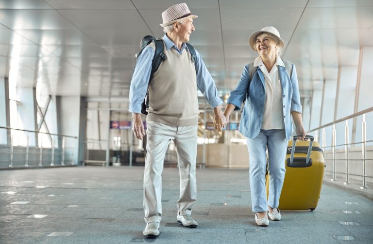 An older couple look adoringly at each other while holding hands and walking through an airport to catch a flight