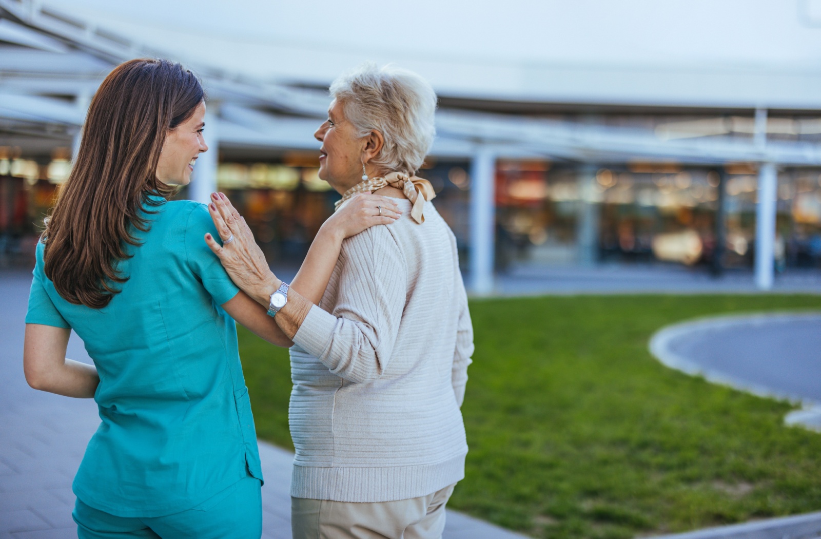 Nurse in teal uniform walking outdoors with smiling senior woman in cream sweater
