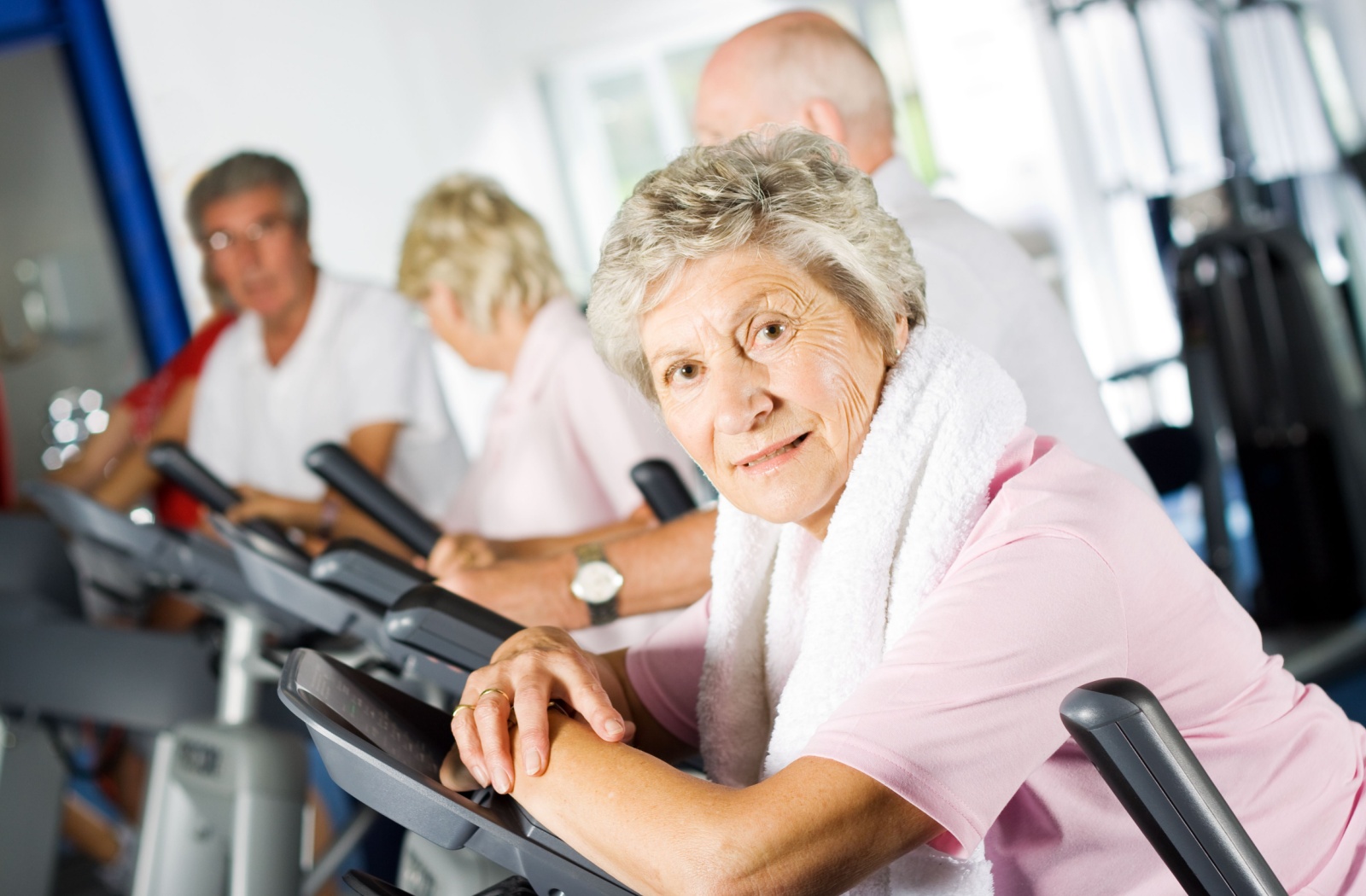 An older adult with a towel around their neck leaning against the display of a treadmill after a cardio fitness class.