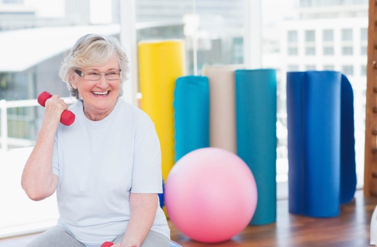 Smiling older adult lifting dumbbells in a bright fitness room with yoga mats and exercise ball for strength training.