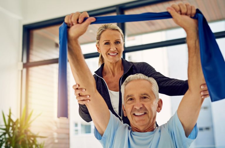 A rehab worker helps a senior stretch their back with a dark blue resistance band in each hand in a bright exercise room