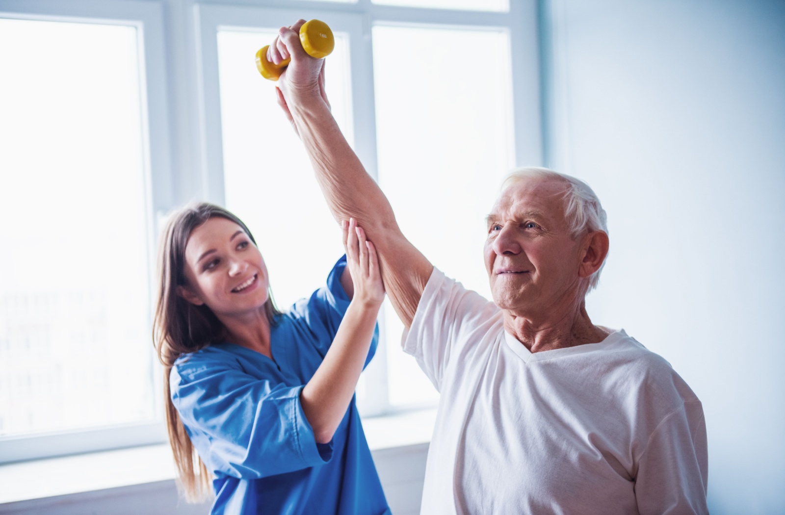 A physical therapist in blue scrubs helps a senior raise a small dumbbell above their head in a bright and white clinical setting
