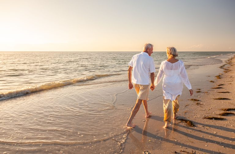 An older couple wearing white linen hold hands while walking along a sandy beach at sunset