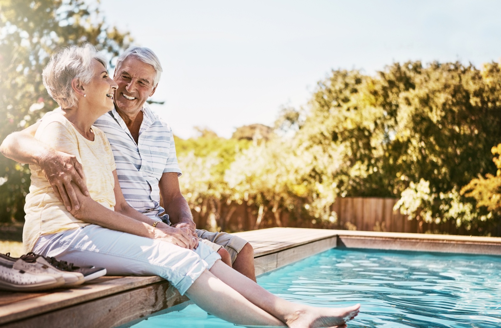 An older adult puts their arm around their spouse's shoulder while they dip their feet in a pool on a summer afternoon