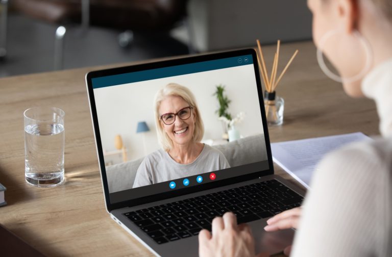 A busy adult child uses a laptop to video call their smiling older parent from their workplace
