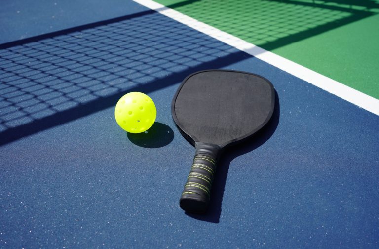 A close-up image of a pickleball paddle and wiffle ball sitting on a blue sunlit outdoor court