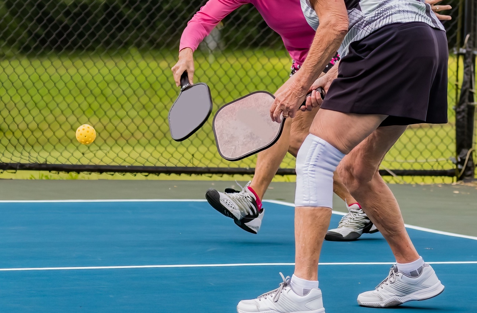 An older adult with a knee brace bends beside their partner to return a mid-air pickleball during a game on an outdoor court
