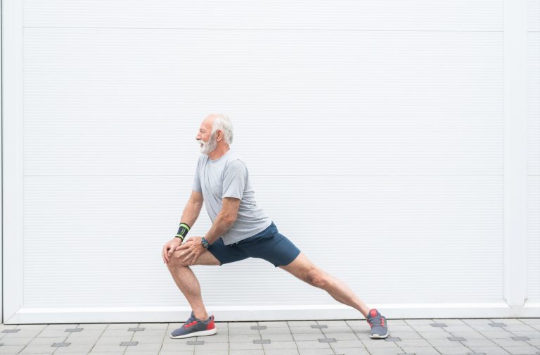 An older adult performs a standing lunge to the left while walking indoors in front of a gray wall