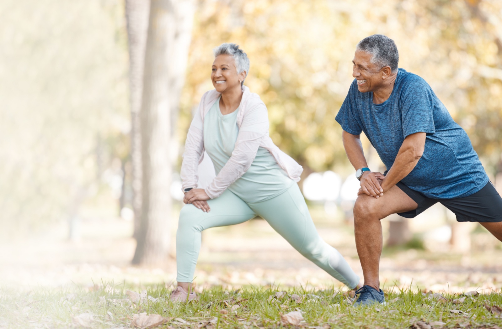 2 older adults stop and perform lunges in the park to stretch and strengthen their legs while out on a walk in the fall

