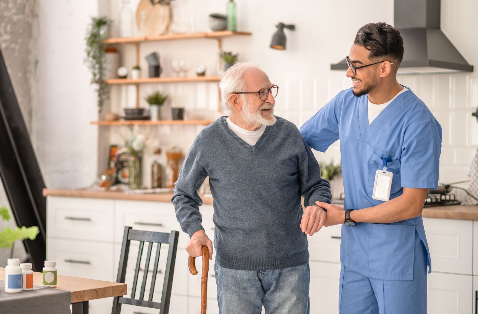 An older adult with a cane smiles while a caregiver helps them stand up from their kitchen table after managing their medication

