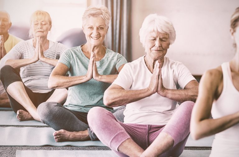 A group of older adults smile while pressing their hands together and stretching cross-legged during a guided yoga meditation.