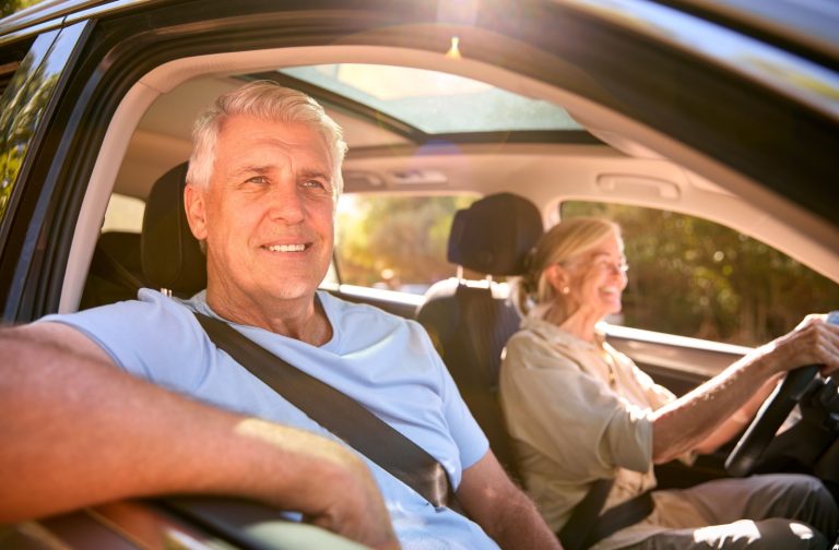 A senior couple enjoys a drive in their car on a sunny day, not worrying about home maintenance since they live in an independent living community.
