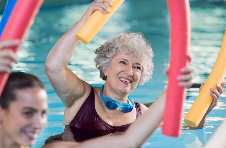 A smiling senior is doing water aerobics with swim noodles during a fitness class in their senior living community.