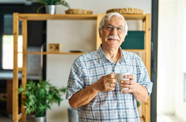 An older adult stands in their kitchen and wraps their hands around a cup of tea while smiling