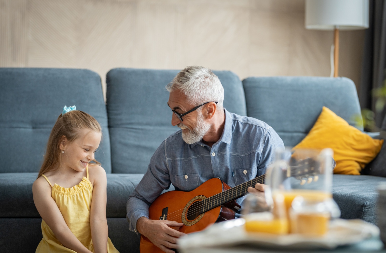 An older adult laughs and sits on the floor with their shy grandaughter with a guitar and tries to get the child to sing