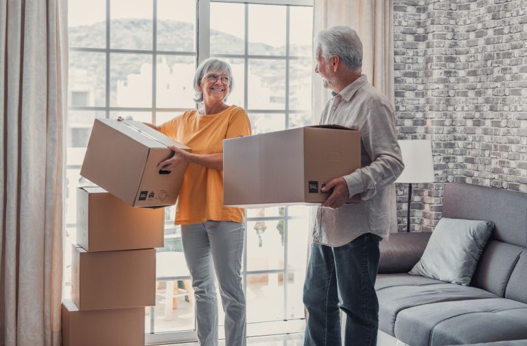 An older couple looks at each other while carrying cardboard boxes in their living room.