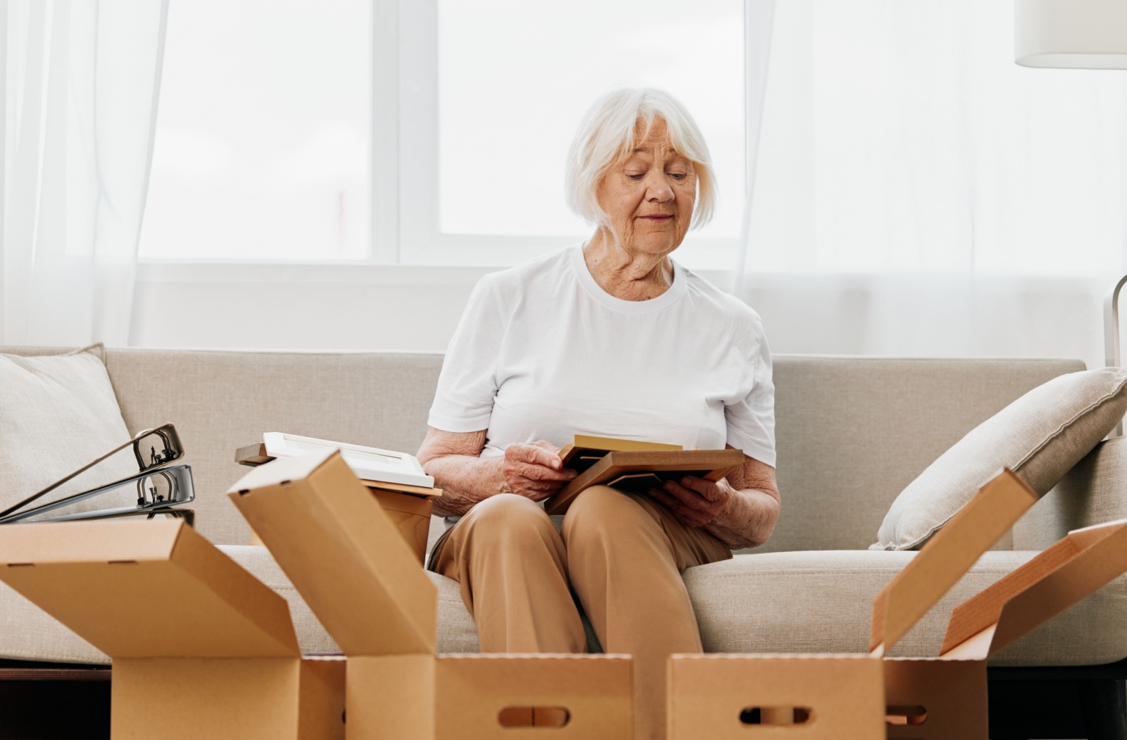 An older adult sits on their couch in front of cardboard moving boxes and sorts through some books.