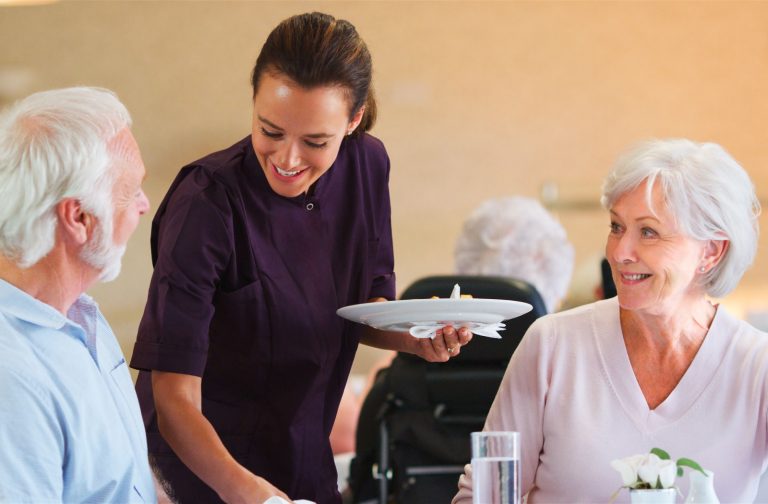 A caregiver picks up plates from 2 smiling residents during mealtime in assisted living