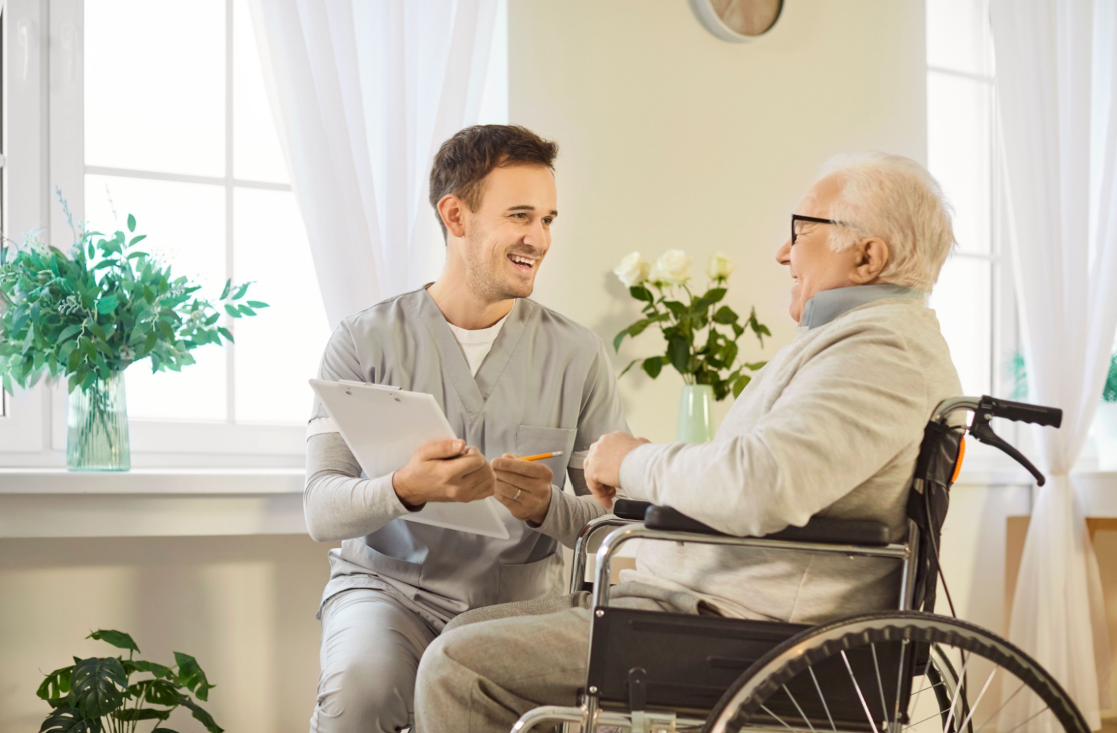 A caregiver holding a clipboard kneels beside a resident and laughs during a quick checkup in assisted living