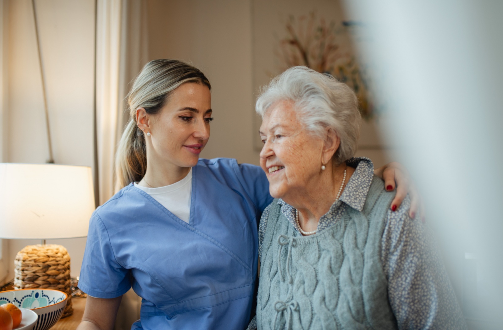 A smiling professional caregiver in light blue scrubs embraces a happy senior with white hair who is wearing a pearl necklace and a knitted vest, in a warm, well-lit indoor setting.