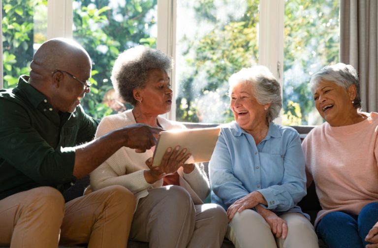 A group of four seniors are sitting together on a couch by a large window, laughing and looking at a tablet being held by one of the individuals.