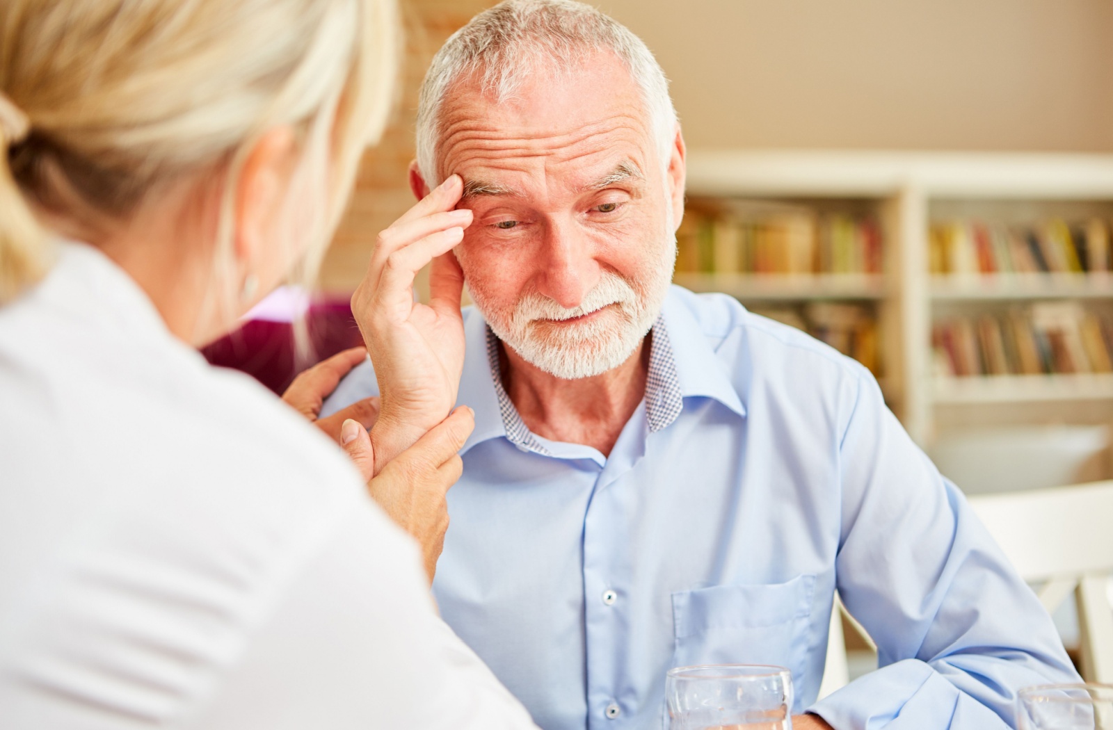 A senior adult with a white beard touches their temple while an adult's hand gently rests on their arm, suggesting comfort or support.
