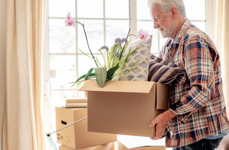 A senior walks in front of a window while carrying a cardboard box full of pillows and an orchid houseplant.