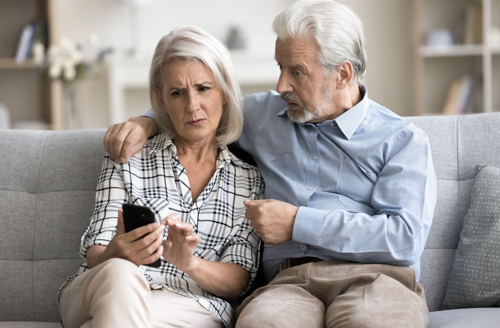 Two senior adults sitting on a couch look concerned while looking at a smartphone.
