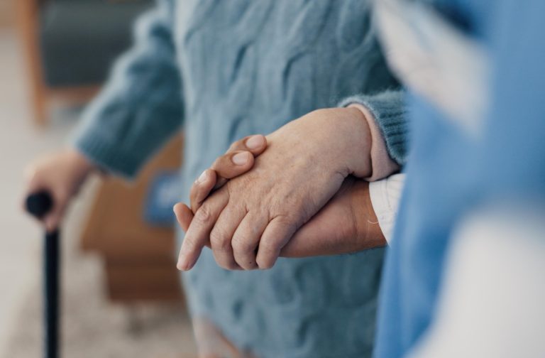 A close-up shot of a caregiver or assistant gently holding the hand of a senior, who is wearing a light blue sweater and holding a cane in the background.