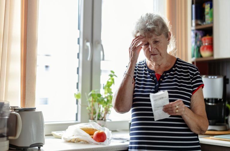 A senior adult in a striped shirt looks distressed while holding a receipt in a kitchen.