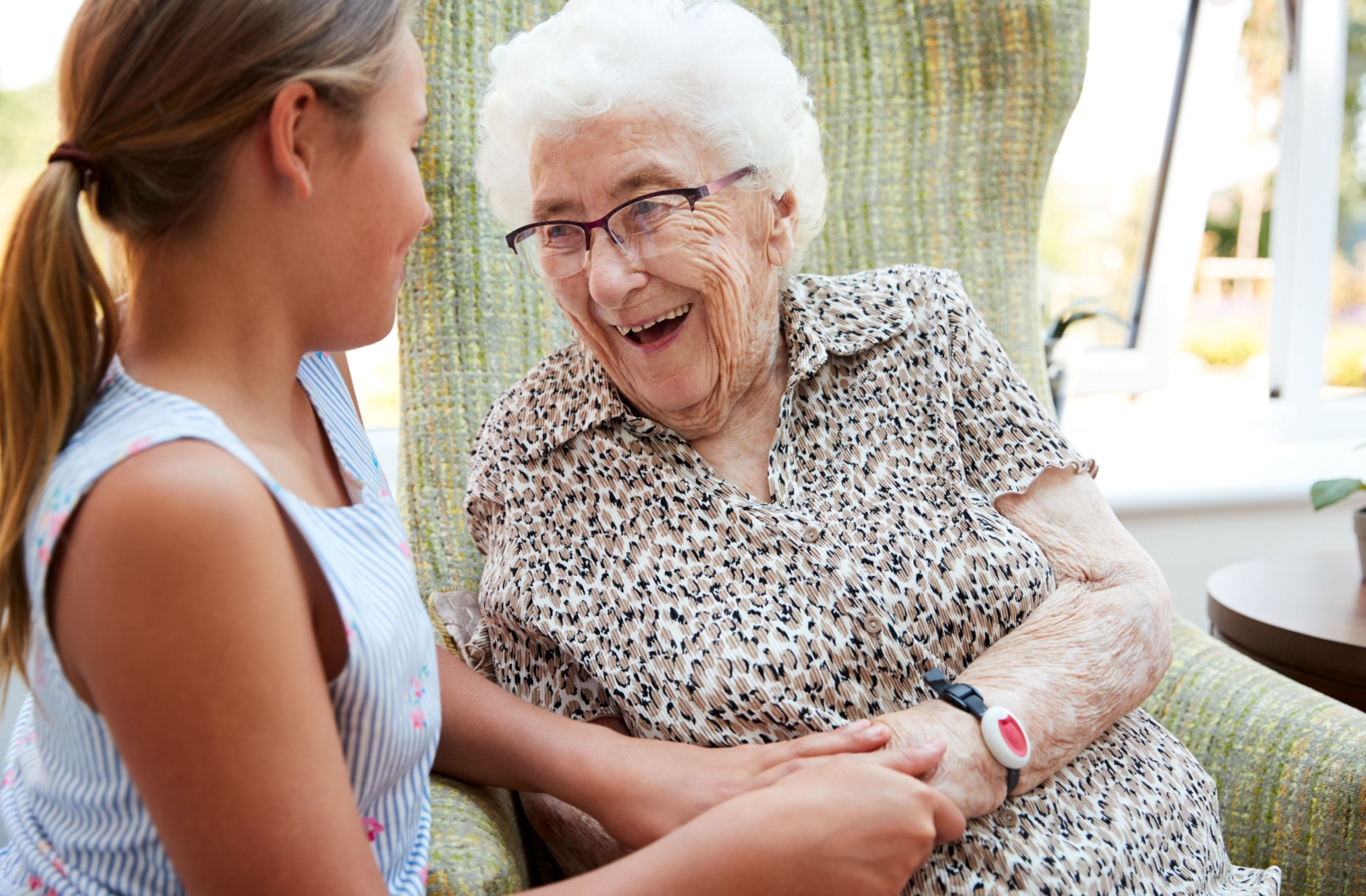 A young person and a senior adult with glasses sit together, holding hands and smiling warmly at each other in a bright setting.
