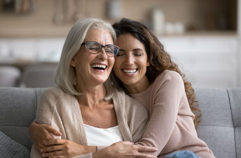 An adult daughter smiles as she hugs her senior mother while they both sit on a gray couch.