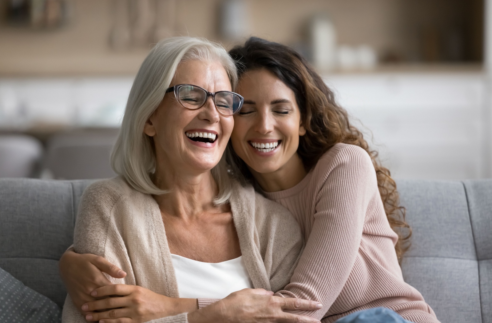 An adult daughter smiles as she hugs her senior mother while they both sit on a gray couch.
